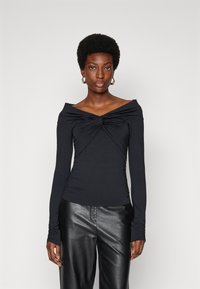 Melnā off-the-shoulder garo piedurkņu blūze ar savītas priekšpuses dizainu, elastīgs audums un saraušanās detaļas pie piedurknēm, apvienota ar melnām ādas biksēm.