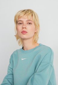 Joven con cabello rubio corto que lleva un sudadera de Nike azul claro, mirando directamente a la cámara frente a un fondo blanco liso.