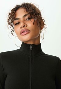 Jeune femme aux cheveux bouclés portant des boucles d'oreilles en or et un haut noir zippé à col montant, fond neutre.