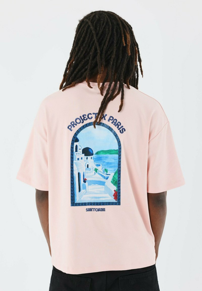 Camiseta de manga corta rosa con una ilustración en azul y blanco de Santorini en la parte posterior, enmarcada con bordes decorativos.