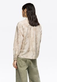 Camisa de manga larga en beige con un patrón de serpiente, con un diseño de pliegue en la parte posterior. Combinada con pantalones sueltos en verde oliva.