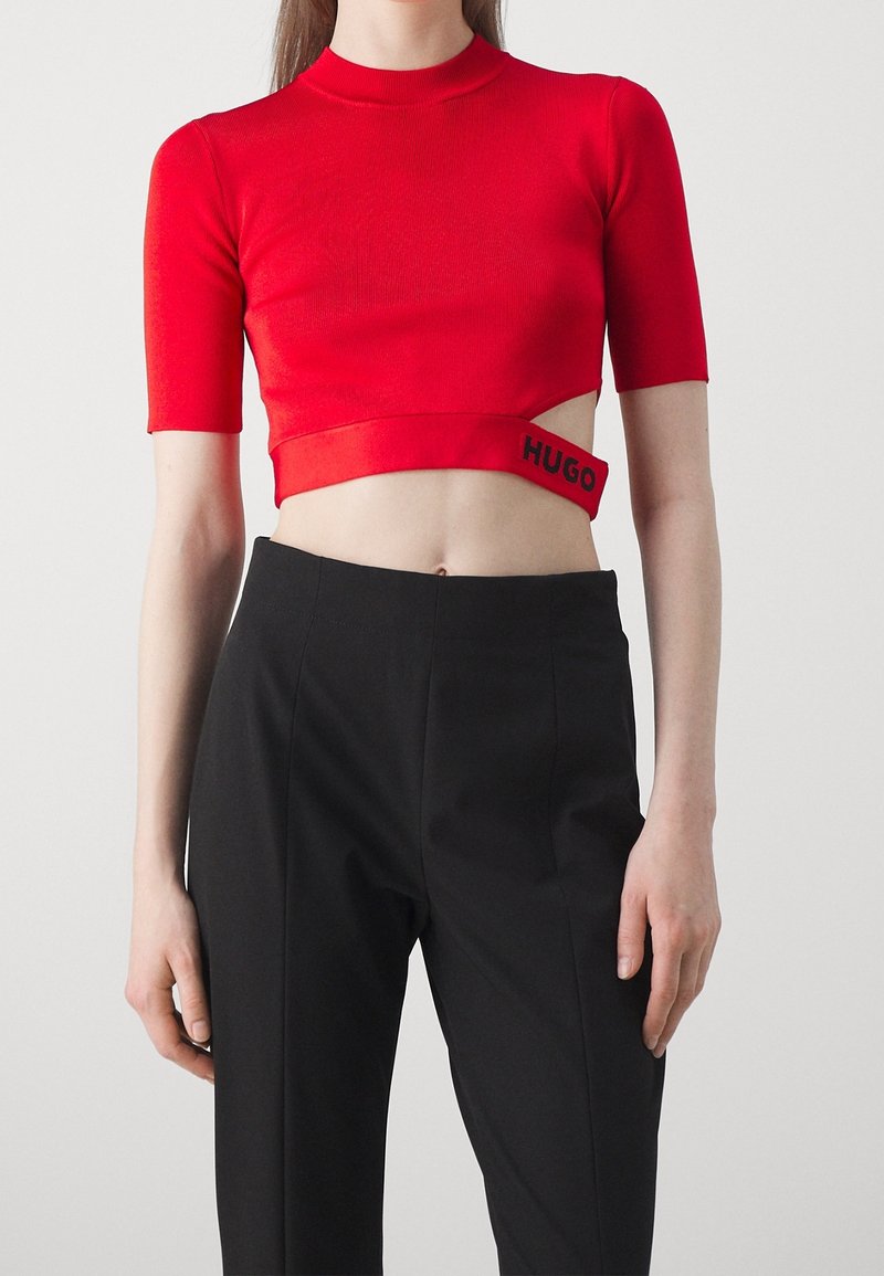 Femme portant un top court rouge avec découpe et logo "HUGO" associé à un pantalon noir taille haute sur un fond clair uni.