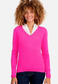 Pull-over rose vif en V, confectionné dans un tissu doux, avec des manches longues et une coupe ajustée, associé à une chemise blanche à col en dessous.