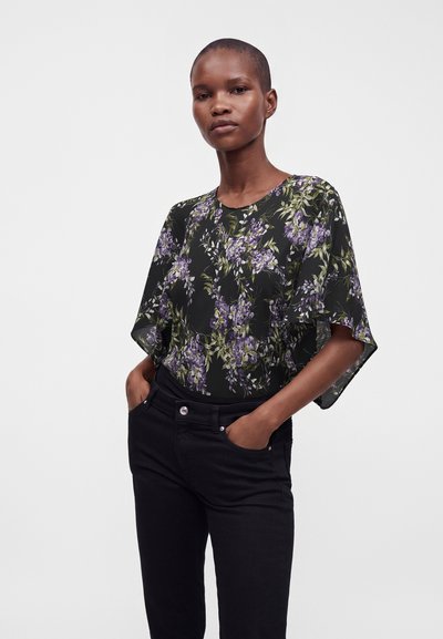 Femme portant un chemisier noir à motifs floraux violets et verts, associé à un pantalon noir, debout avec les mains dans les poches sur un fond uni.