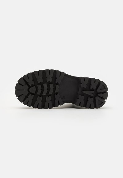 Suela de bota de goma negra con surcos profundos y una superficie texturizada, que presenta patrones angulares para tracción y estabilidad.