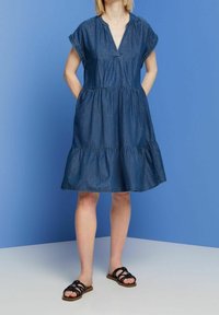 Denim dress with short sleeves, V-neckline, gathered waist, and tiered skirt. Worn with black strappy sandals on a light surface.