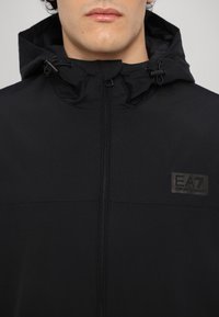 Svart huva jacka med dragkedja framtill, som har ett subtilt texturerat tyg och en tonad EA7 Emporio Armani-logotyp på över vänster sida.