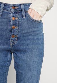 Los jeans de mezclilla azul de cintura alta cuentan con un cierre de cinco botones, dos bolsillos laterales y una textura suave con costuras visibles.