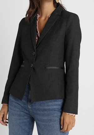 Femme portant un blazer noir cintré à deux boutons sur un chemisier rose à pois et un jean bleu, vue du cou jusqu’aux hanches.