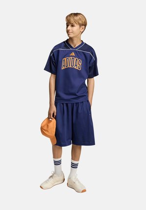 Adolescente con ropa deportiva azul de Adidas con pantalones cortos y camiseta, sosteniendo una gorra naranja en una mano, zapatillas blancas y calcetines a rayas.