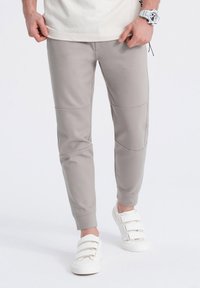 Pantalones jogger de color gris claro en una tela suave; diseño ajustado con detalles de paneles en las rodillas. Combinados con zapatillas blancas de Velcro.