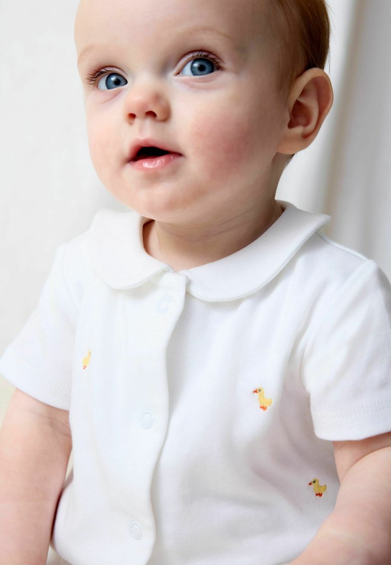 Bébé aux yeux bleus et aux cheveux clairs portant une chemise blanche à col décorée de petites illustrations de canards jaunes.