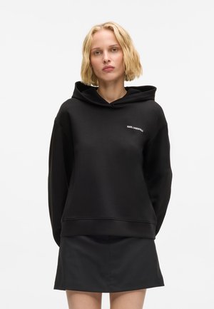 Lys hård kvinde med kort hår iført en sort Karl Lagerfeld hættetrøje og sort nederdel, vendende fremad mod en ensfarvet hvid baggrund.