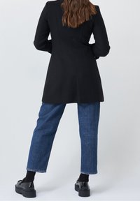 Cappotto nero con design strutturato, maniche lunghe e cuciture verticali. Abbinato a jeans in denim blu e scarpe con zeppa nere.