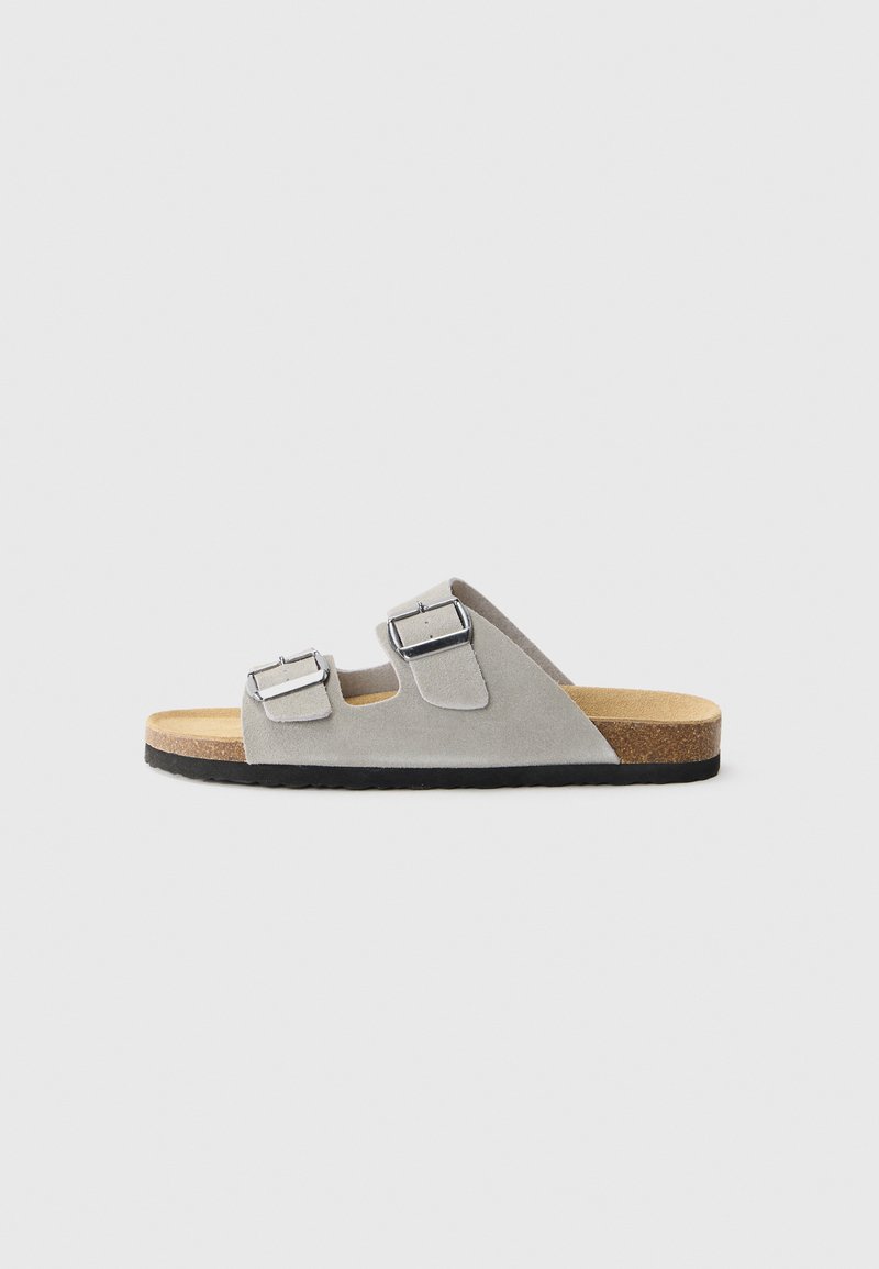 Sandal din suede gri, cu talpă slide, prevăzută cu două catarame argintii ajustabile, un suport din plută, o talpă din cauciuc texturat negru și o formă rotunjită pentru confort.
