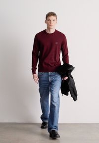 Μπορντό πλεκτό πουλόβερ με τετράγωνη λεπτομέρεια, ανοιχτό μπλε τζιν και μαύρα παπούτσια, κρατώντας ένα μαύρο μπουφάν, περπατώντας σε ουδέτερο φόντο.