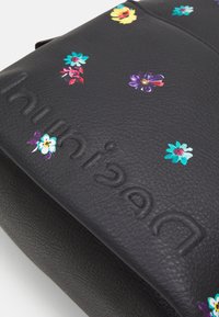 Černá texturovaná kožená taška s malými barevnými květinovými vzory a razítkovaným názvem značky na povrchu.