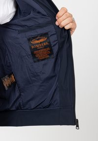 Navy-Bomberjacken-Innenfutter mit Satinfinish, ausgestattet mit einem orangefarbenen Tag, der "BOMBERS ORIGINAL" beschriftet ist, sowie Pflegeetiketten auf der linken Seite.