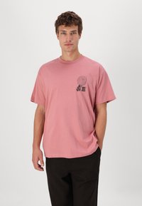 Camiseta de algodón de manga corta en color rosa, con un diseño gráfico negro en el lado izquierdo del pecho. Cuello redondo clásico y ajuste relajado.