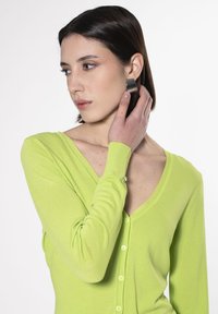 Cardigan verde lime in tessuto morbido con scollo a V, maniche lunghe e chiusura con bottoni; modello tiene un orecchino a cerchio argentato vicino all'orecchio.