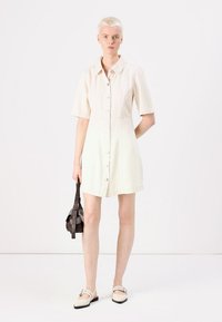 Robe à manches courtes à boutons crème, avec un corsage ajusté et une jupe évasée. Portée avec un petit sac sombre et des chaussures légères.