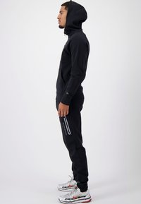 Zwarte capuchontrui en bijpassende joggingsbroek. Glad materiaal, slim fit, grijze accenten op de broek. Model draagt witte en rode sneakers.