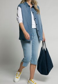 Denim sleeveless shirt in light blue, buttoned down with a collar, paired with light blue capris and white sneakers with yellow accents.