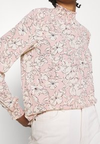 Blommig blus i ljusrosa med svart och vit blommönster. Har en hög volangkrage och rynkade manschetter på lång ärm.