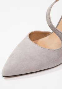 Chaussure plate en daim gris clair à bout pointu avec une fine bride à la cheville et une doublure intérieure en cuir beige sur fond blanc.