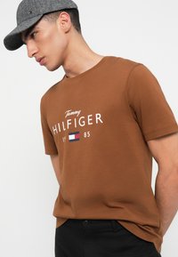 Muž ve hnědém tričku Tommy Hilfiger a šedé herringbone kšiltovce, pózující před jednoduchým bílým pozadím.