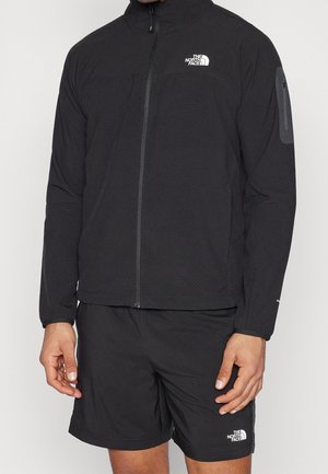 Homme portant une veste noire The North Face zippée entièrement avec un logo sur la poitrine et un short noir avec un logo sur la cuisse gauche, debout, mains détendues.