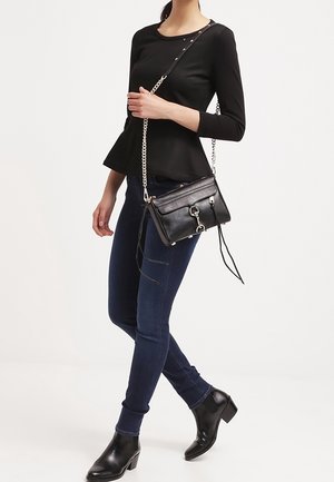 Zwarte crossbodytas met kettingriem, getextureerd leer, ritssluiting en tasselaccenten. Draagt bij een strakke jeans en enkellaarzen.