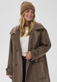 Hnědý vlněný kabát s velkým límcem, dvojitými knoflíky a přední kapsou. Nošený přes lehký svetr a kombinovaný s hnědou čepicí.