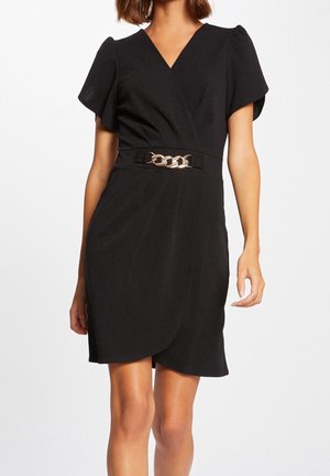 Schwarzes Wickelkleid mit kurzen Flatterärmeln und einem Taillengürtel mit goldener Kettendetail, getragen von einer stehenden Person.