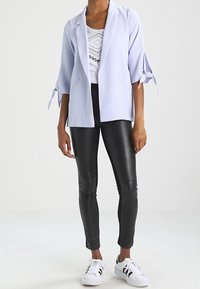 Blazer bleu clair à manches trois-quarts avec poignets noués, porté sur un haut blanc graphique, associé à des leggings noirs et des baskets blanches.