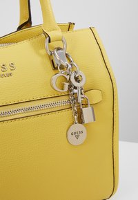 Keltainen tekstuuri nahkalaukku, jossa on hopeinen vetoketju ja metallisia avaimenperikoruja, mukaan lukien lukko, avain, G-kirjain ja pyöreä Guess-logo-tag.