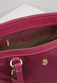 Ciemnoróżowa torebka z złotym logo U.S. Polo Assn., widoczną kieszonką na zamek błyskawiczny w środku oraz białymi szwami wzdłuż górnej krawędzi.