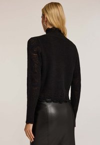 Svart stickad tröja med hög krage, texturerade ärmar, scallop-kant och ett ribbat mönster. Kombinerad med en slimmad svart läderskjorta.