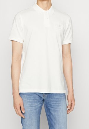 Mężczyzna ubrany w gładką, białą, krótko rękawową koszulkę polo z dwoma guzikami oraz jasnoniebieskie dżinsy, stojący na białym tle.