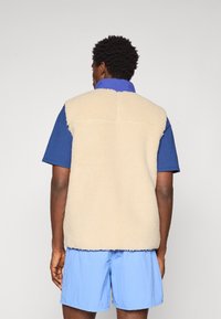 Beige fleece vest with a soft texture, featuring a high collar and no sleeves. Worn over a blue shirt and paired with light blue shorts.