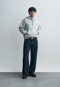 Φωτεινό γκρι φούτερ με μπροστινή τσέπη και ρυθμιζόμενο κουκούλι, συνδυασμένο με σκούρο μπλε τζιν με φαρδιά μπατζάκια. Μαύρα παπούτσια ολοκληρώνουν το καθημερινό look.