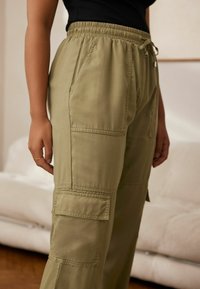 Helle khaki Cargo-Hosen mit einem elastischen Bund, seitlichen Taschen und gestickten Akzenten. Das Material wirkt glatt und hat eine lockere Passform.
