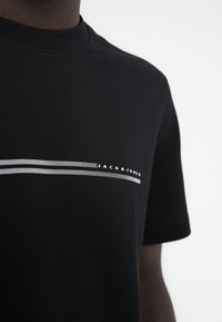 Μαύρο βαμβακερό T-shirt με οριζόντια λευκή ριγέ σχεδίαση και κείμενο «JACK & JONES». Κοντά μανίκια, στρογγυλή λαιμόκοψη.
