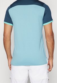 Polo deportivo de color azul claro con paneles en el hombro en navy y acentos en turquesa. Mangas cortas, tejido texturizado, aberturas laterales en el dobladillo.