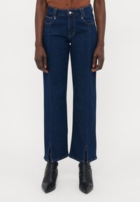 Jeans de perna larga em azul-escuro, feitos de denim, com bolsos frontais, fecho de botão e fendas laterais distintas nos tornozelos.