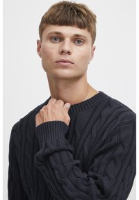 Dunkelblauer Strickpullover mit strukturiertem Kabelmuster, geripptem Rundhalsausschnitt und Bündchen. Der Stoff wirkt dick und warm.