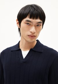 Marineblauwe gebreide trui met verticale ribbels, v-hals en kraag, met een zachte textuur en een aansluitend ontwerp. Gedragen over een wit shirt.