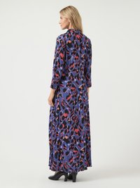 Egy szőke hajú nő hosszú, lila ruhát visel, amelyen piros és fekete absztrakt minták találhatók, fekete magassarkú cipőben áll egy fehér háttér előtt.