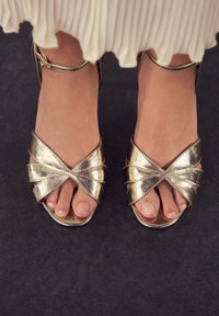 Metallic goldene Sandalen mit offenem Zehenbereich, überkreuzten Riemen, glatter Oberfläche und Knöchelriemen. Zeigt nackte Zehen und klare Nagellackierung.