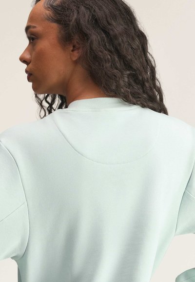 Kobieta o kręconych włosach, ubrana w jasnoniebieską bluzę, zwrócona bokiem, prezentująca tył i szczegóły ramienia ubrania.